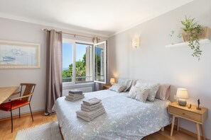 Ein Schlafzimmer mit Bett, Schreibtisch, Stuhl, einem Fenster mit Vorhängen, einem Nachttisch mit Lampe und einem an der Wand befestigten Regal mit Pflanze.