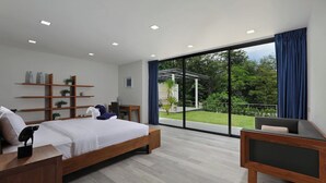 Ein modernes Schlafzimmer mit einem großen Bett, einem Schreibtisch, einem Stuhl und einem Fernseher. Eine Glastür führt zu einem Balkon mit Blick auf Grünflächen.