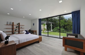 A modern bedroom with a large bed, a desk, a chair, and a TV. There is a sliding glass door leading to a balcony with a view of greenery. - Sugar | Kamala Beach | 4-bedroom Pool Villa (Kamala)