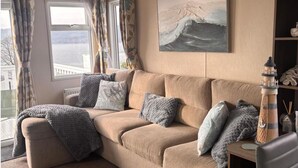 A living room with a beige sofa, patterned cushions, a painting of a wave, and a lighthouse model. - Seaside Escape in Wemyss Bay (Wemyss Bay)