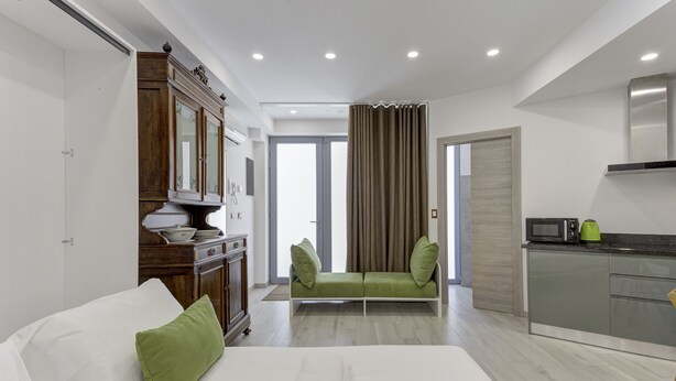 A modern hotel room with a bed, green pillows, a wooden cabinet, a green sofa, a kitchen area, and a view through a window. - Varenna Garden by Wonderful Italy (Perledo)