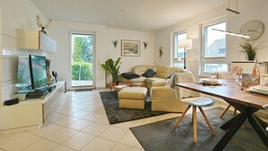 A modern living room with a sofa, coffee table, and dining table. There is a television, a potted plant, and a window providing natural light. - Home port apartment 1 (Scharbeutz)