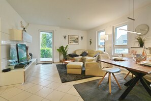 Ein modernes Wohnzimmer mit Sofa, Couchtisch und Esstisch. Ein Fernseher, eine Zimmerpflanze und ein Fenster, das natürliches Licht hereinlässt, sind ebenfalls vorhanden.