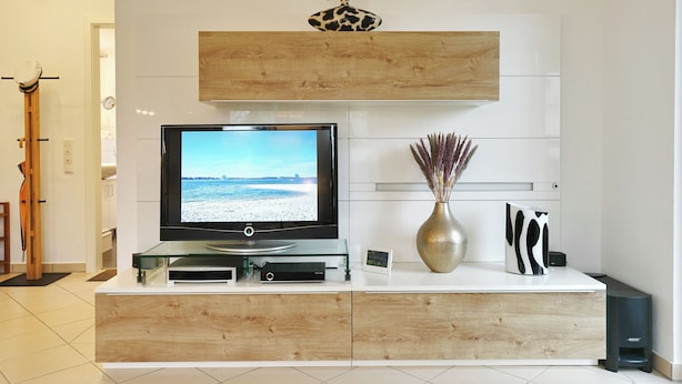 A modern living room with a flat-screen TV, wooden cabinet, decorative vase, and a mounted shelf. - Home port apartment 1 (Scharbeutz)