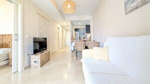 Un salon moderne avec un canapé blanc, un téléviseur, une décoration murale en macramé et, au fond, un coin repas.