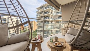 Un balcon avec des fauteuils en osier et une petite table, une vue sur un immeuble à plusieurs étages et un ciel dégagé.