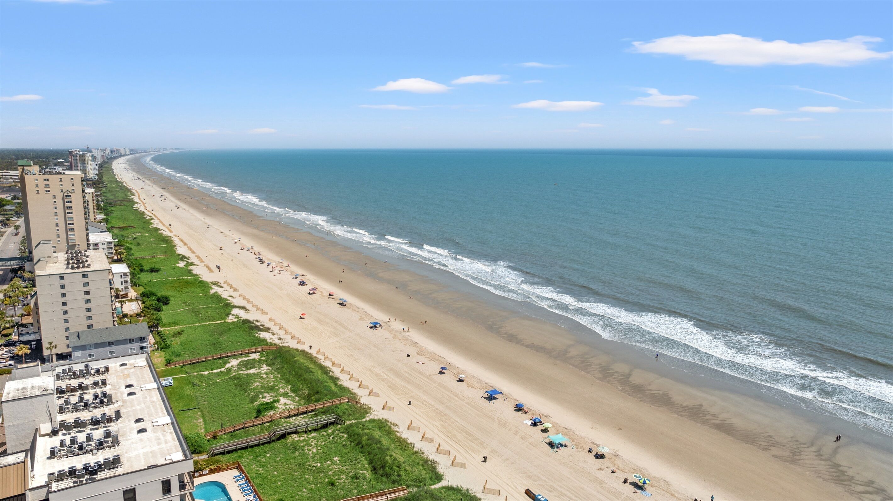 A coastal beach with a long stretch of sand, a few umbrellas, and a building with a rooftop pool.
