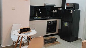 Een compacte keuken met witte kasten, een zwarte koelkast, een magnetron en een gasfornuis. Er is een klein eethoekje met een witte tafel en twee stoelen. Aan de muur hangt een schilderij van een zonsondergang.