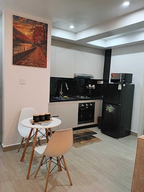 A compact kitchen with white cabinets, a black refrigerator, a microwave, and a gas stove. There is a small dining area with a white table and two chairs. A painting of a sunset is on the wall. - 1 bedroom  kilimani in Nairobi (Nairobi)