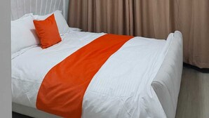 Een hotelkamer met een bed, wit beddengoed, een oranje sierkussen en een groot raam met gordijnen.