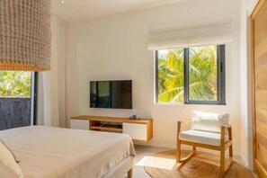 Ein Schlafzimmer mit Bett, Fernseher, Sessel und Fenster mit Blick auf Palmen.