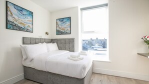 A modern bedroom with a grey bed, white bedding, two abstract paintings, a window with a city view, and a small vase with flowers.