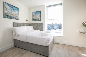 A modern bedroom with a grey bed, white bedding, two abstract paintings, a window with a city view, and a small vase with flowers. - Portobello Court Apartments (Dublin)