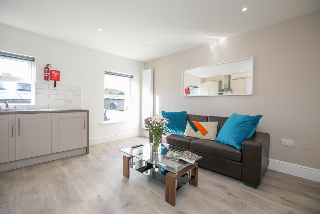 A modern living room with a glass coffee table, a brown sofa with blue cushions, a kitchen area with white cabinets, and a large mirror on the wall.