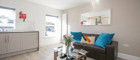 A modern living room with a glass coffee table, a brown sofa with blue cushions, a kitchen area with white cabinets, and a large mirror on the wall.