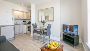 A modern kitchen with white cabinets, a dining area with a table and chairs, a television, and a fruit bowl on a coffee table.