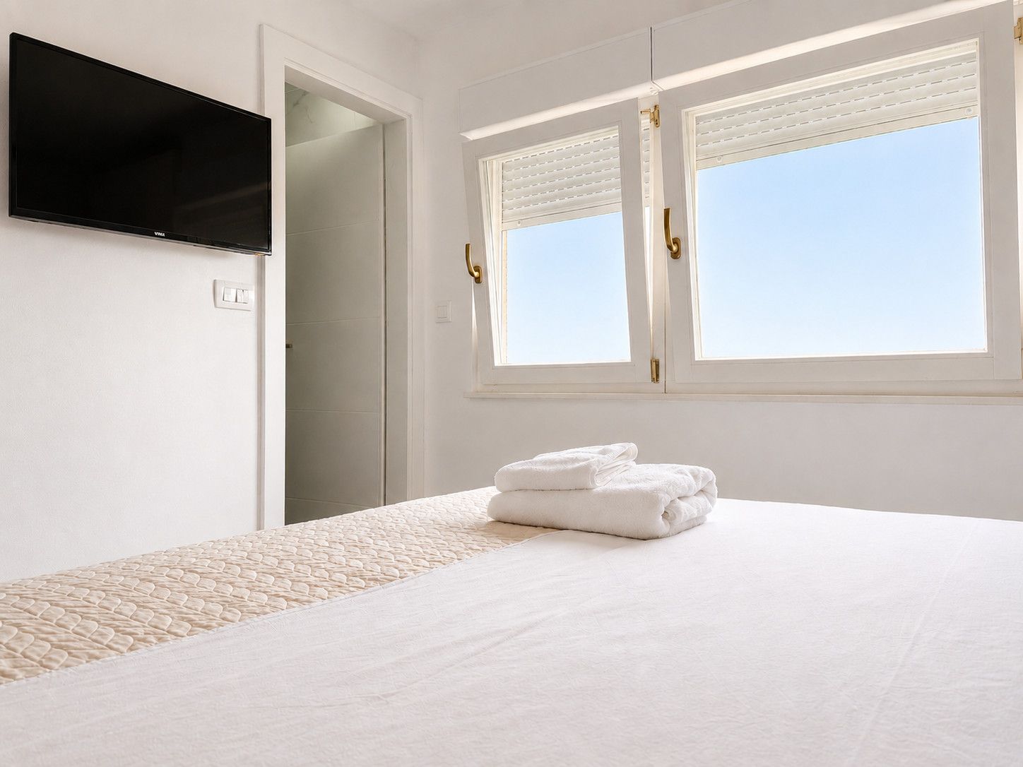 A hotel room with a bed, a flat-screen TV, a window with blinds, and a door.