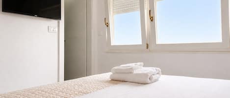 A hotel room with a bed, a flat-screen TV, a window with blinds, and a door.