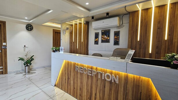 A reception area with a wooden reception desk, a clock, and framed artwork on the wall. - HOTEL BELVA (Ahmedabad)