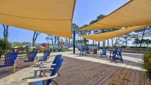 Een houten terras met blauwe stoelen en beige zonneschermen, met uitzicht op een strand met kleurrijke boten.