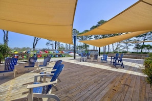 A wooden deck with blue chairs and tan canopies, overlooking a beach with colorful boats.