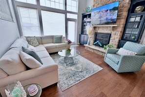 A living room with a beige sofa, a patterned rug, a glass coffee table, a blue armchair, a fireplace, and a TV mounted on the wall.