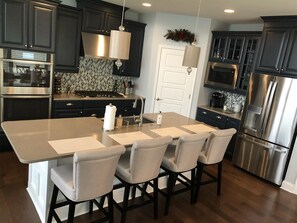 A modern kitchen with dark cabinetry, stainless steel appliances, a central island with chairs, and a hanging light fixture.