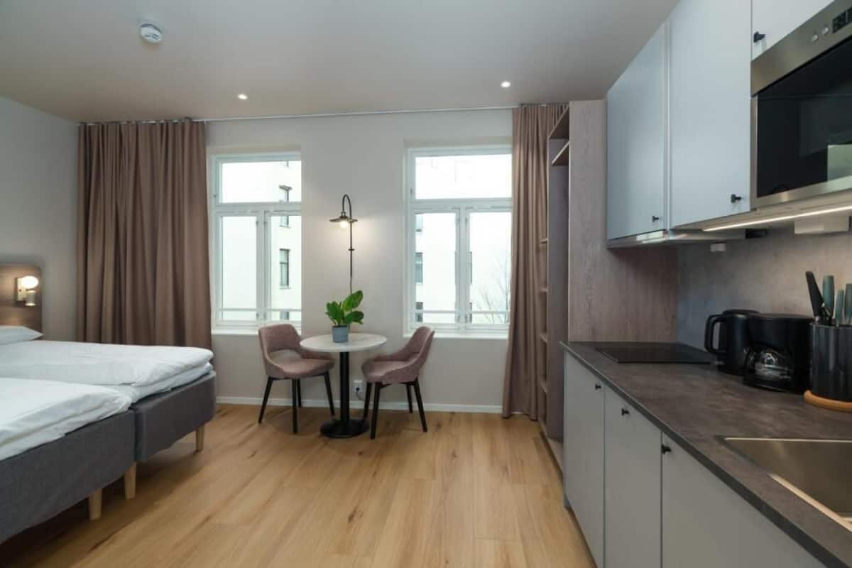 A modern hotel room with a bed, a small dining table with chairs, a potted plant, a large window, a kitchenette with a microwave, and a coffee maker.