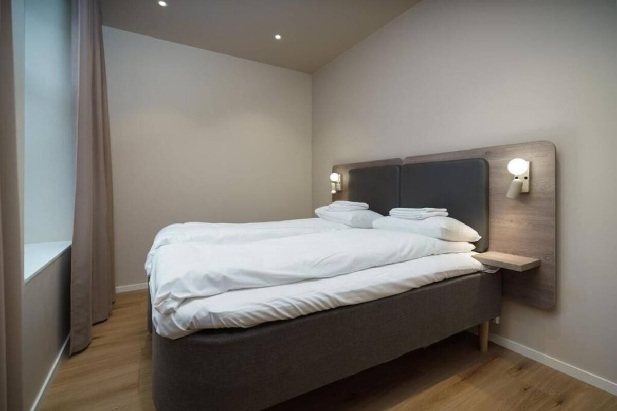 A neatly made bed in a hotel room with a headboard, bedside lamps, and a window with curtains.