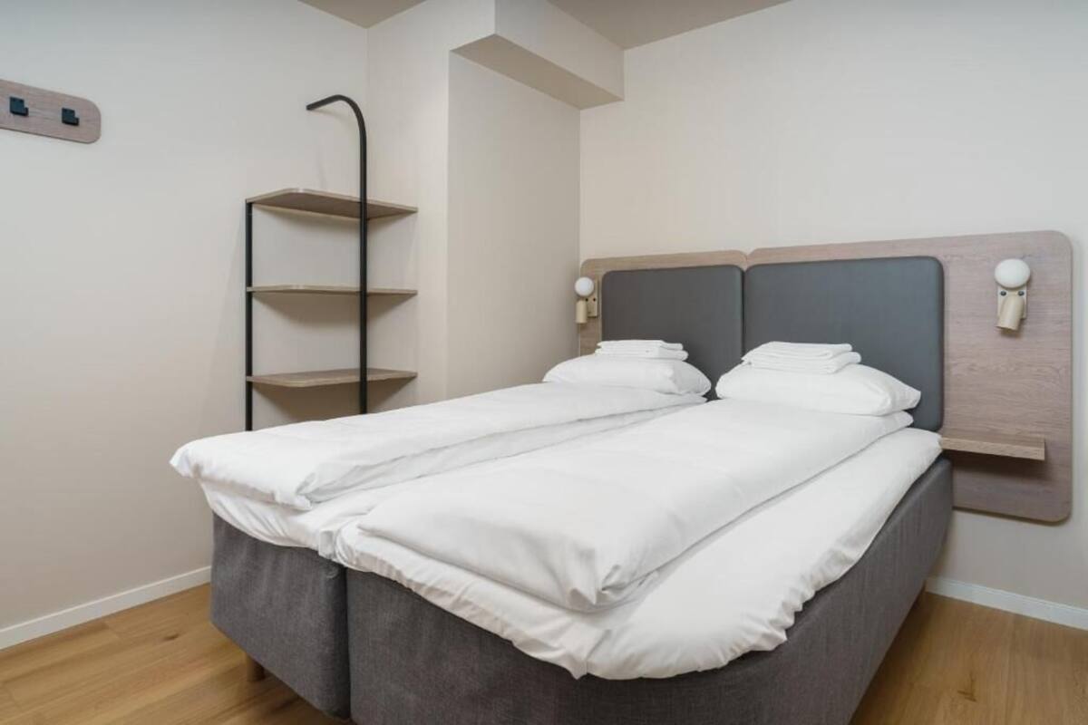 A bed with a grey headboard, white bedding, and a wooden shelf unit beside it.