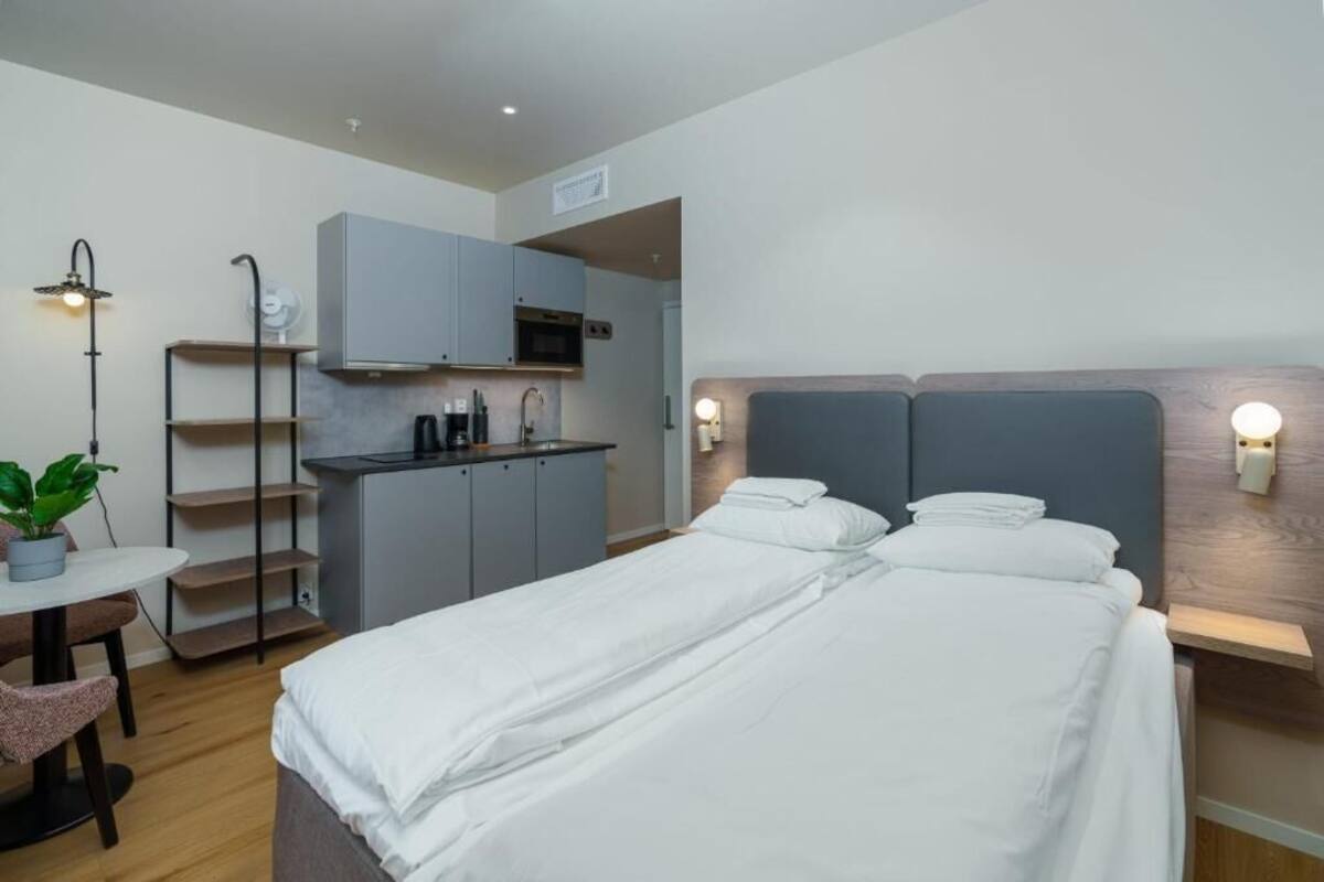 A compact hotel room with a bed, bedside tables, a small kitchenette, and a potted plant.