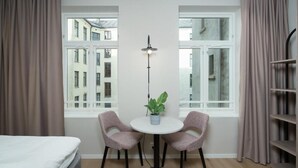 Una habitación con una cama, una mesa redonda con dos sillas, una planta en maceta, una lámpara de pie y vistas a edificios a través de ventanas grandes.