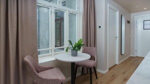 Una habitación con una mesa redonda, dos sillas, una planta en maceta, una ventana con cortinas y una puerta.