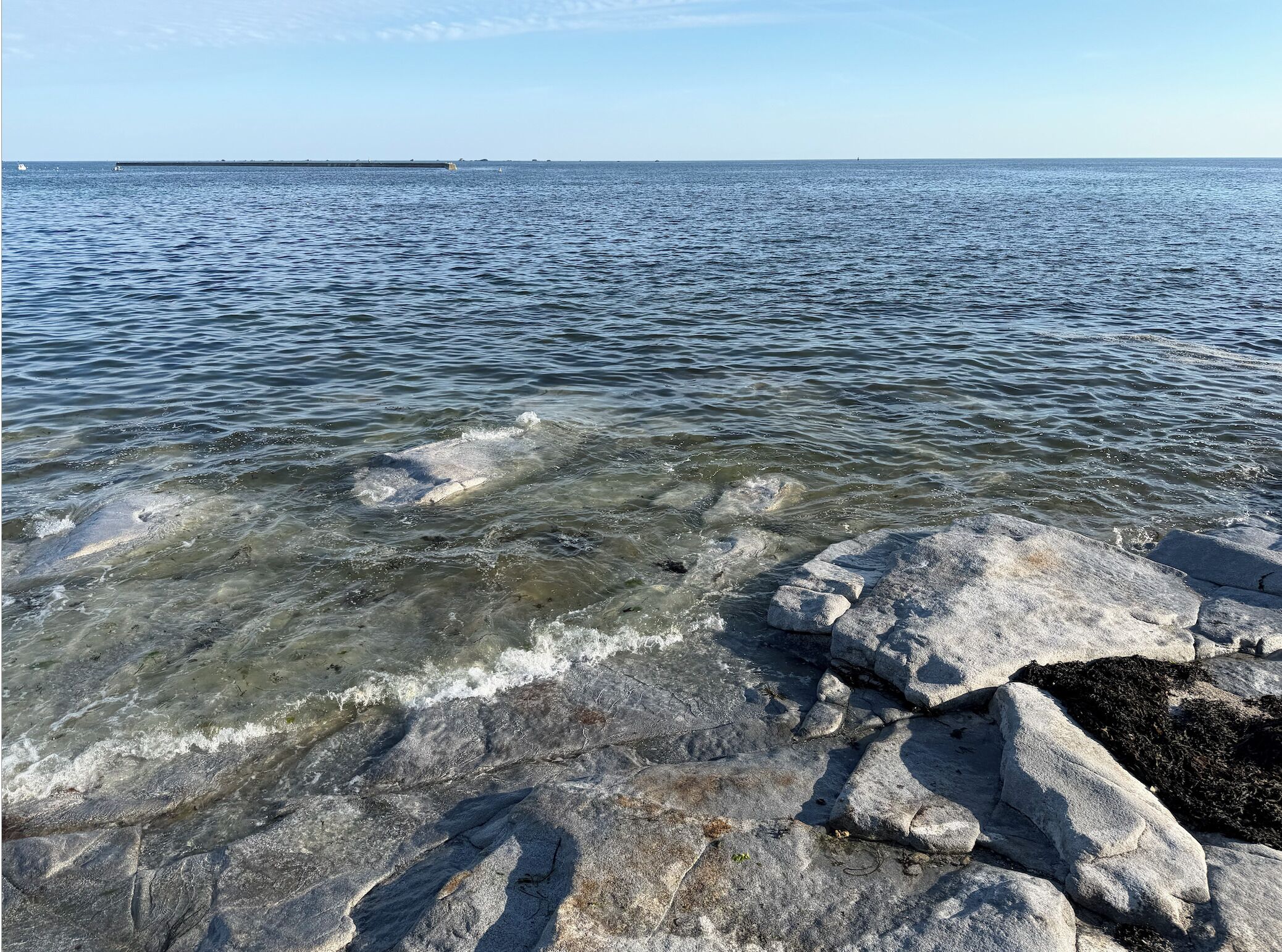 A rocky shoreline with clear water and a distant pier.