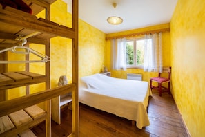 A room with a bed, wooden floor, a window with curtains, a chair, and a hanging light fixture.