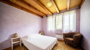 A bedroom with a bed, a chair, a nightstand, a lamp, and a window with curtains. - Cool Nature Retreat with Pool in the Heart of the Forest (Le Soulié)