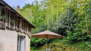 A rustic wooden building with a gravel patio, a picnic table with an umbrella, and a small table with a chair. - Cool Nature Retreat with Pool in the Heart of the Forest (Le Soulié)