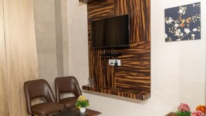 A room with two brown leather chairs, a small table with a vase of flowers, a mounted TV, and a wooden accent wall. - Rosewood By Zenvana (Dehradun)