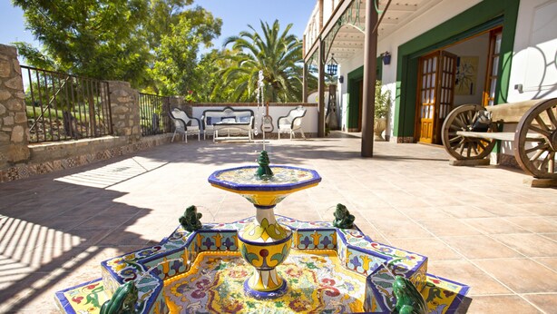 Un patio con una fuente colorida, mesas al aire libre y la entrada de un edificio.