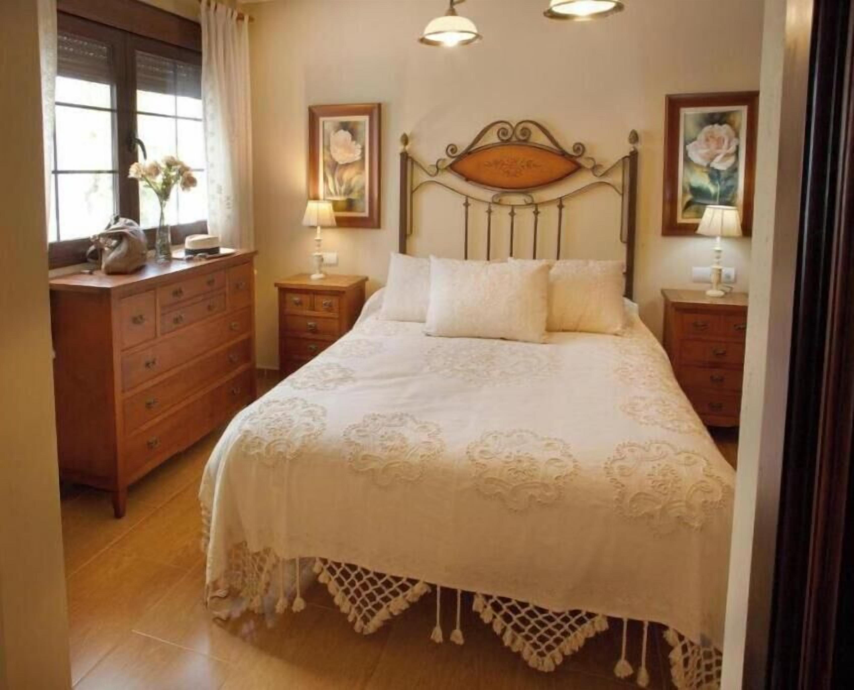 A bedroom with a wooden headboard, a bed with white linens, two bedside tables with lamps, a dresser, and a window with curtains.