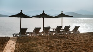 Una platja amb gandules i ombrel·les, una passarel·la de fusta i vista al mar i a les muntanyes llunyanes.