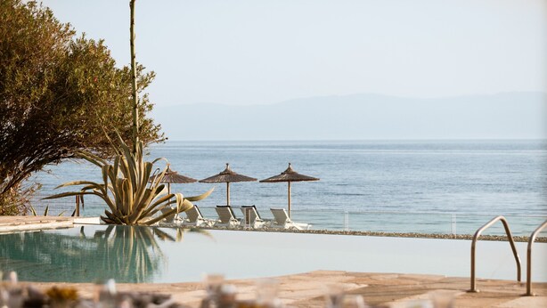 Vista a la piscina amb gandules i ombrel·les, cel blau i una vista llunyana del mar.