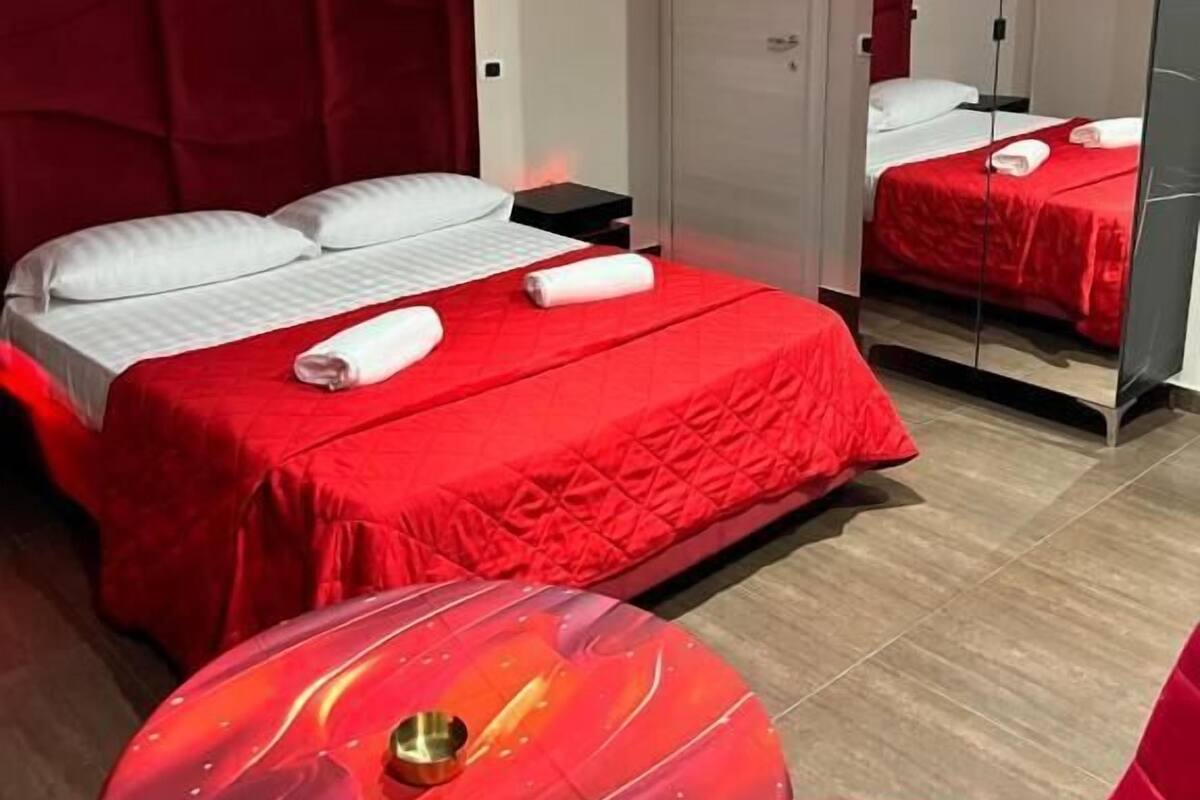 Una habitación de hotel con una colcha roja, un cabecero rojo, una mesita de noche, un espejo y un sofá rojo.