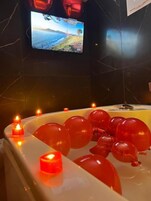 Una bañera con globos rojos y velas, un televisor en la pared, y un piso de baldosas oscuras.