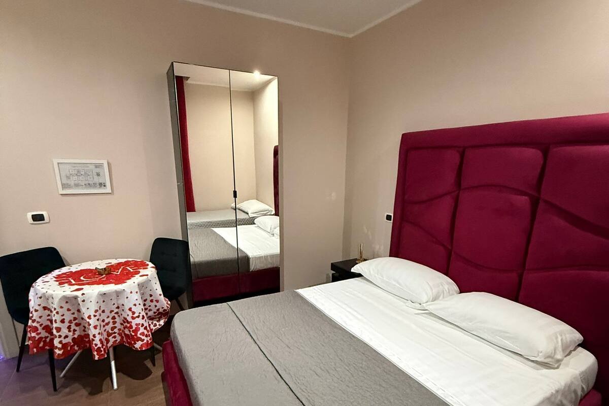 Un dormitorio con una cama, una mesa redonda con mantel rojo y blanco, una silla, un espejo y un cabecero rojo.