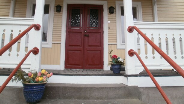 Ein zweistöckiges Haus mit roter Tür, weißen Geländern und einem blauen Blumentopf vor der Haustür.
