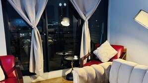 Quarto com sofá de couro vermelho, mesa redonda pequena com luminária e vista para o horizonte urbano através de grandes janelas com cortinas.
