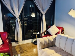 A room with a red leather sofa, a small round table with a lamp, and a view of a cityscape through large windows with curtains. - Home and away from home with a city view. (Johannesburg)