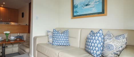 Una sala de estar con un sofá color beige, cojines estampados, un cuadro enmarcado y una cocina al fondo.