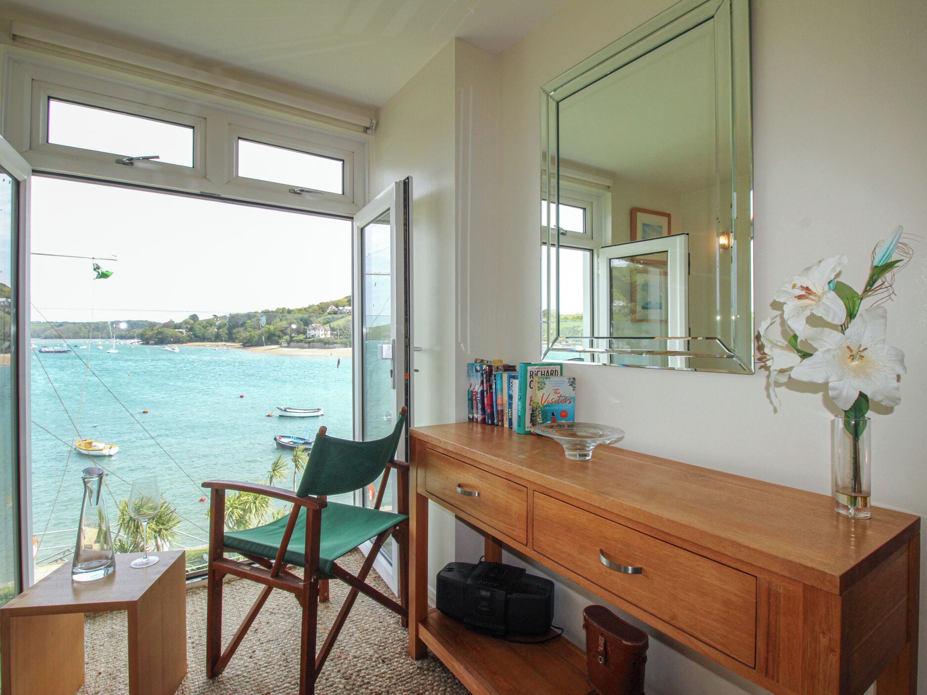 A room with a view of the sea, a chair, a wooden desk, a mirror, and a vase with flowers.